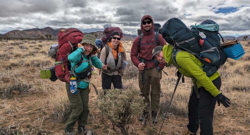 A group of people wearing large backpacks stand smiling in a desert environment. 